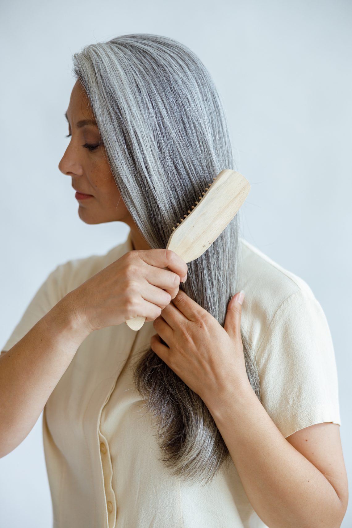 Languid middle aged Asian woman brushes natural hoary hair on light grey background