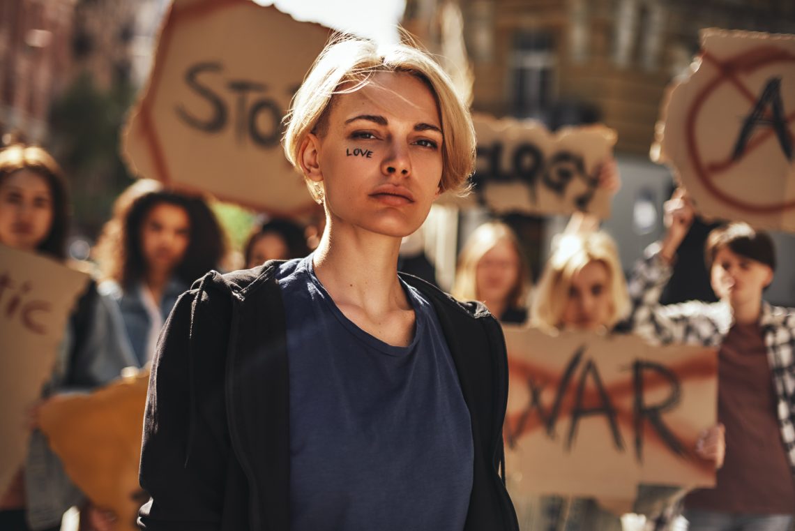 Make love. Young blonde woman with word love written on her face protesting with group of female activists outdoors on road.