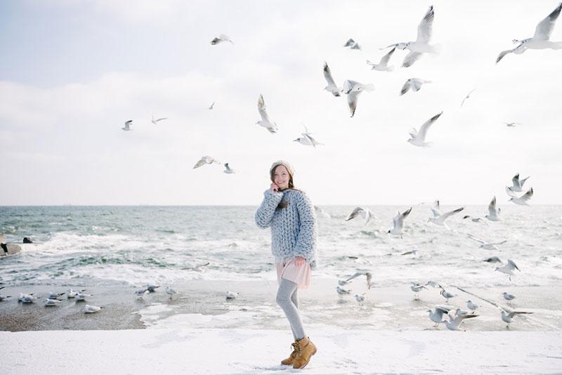 Young woman in merino wool sweater on winter seashore with seagulls