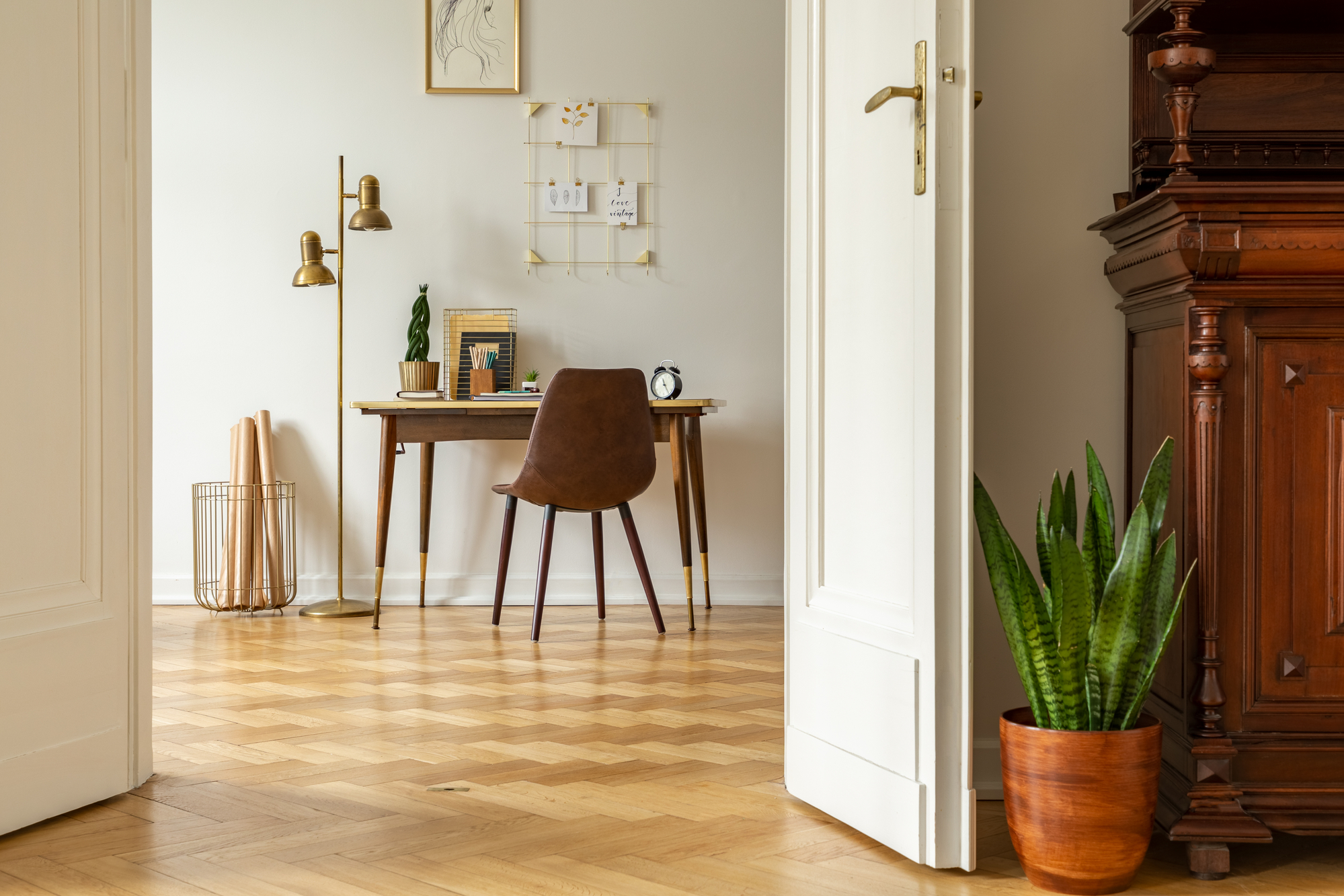 Planta verde por porta branca no interior do escritório de casa de um freelancer com lâmpada de assoalho industrial dourada por uma mesa de madeira