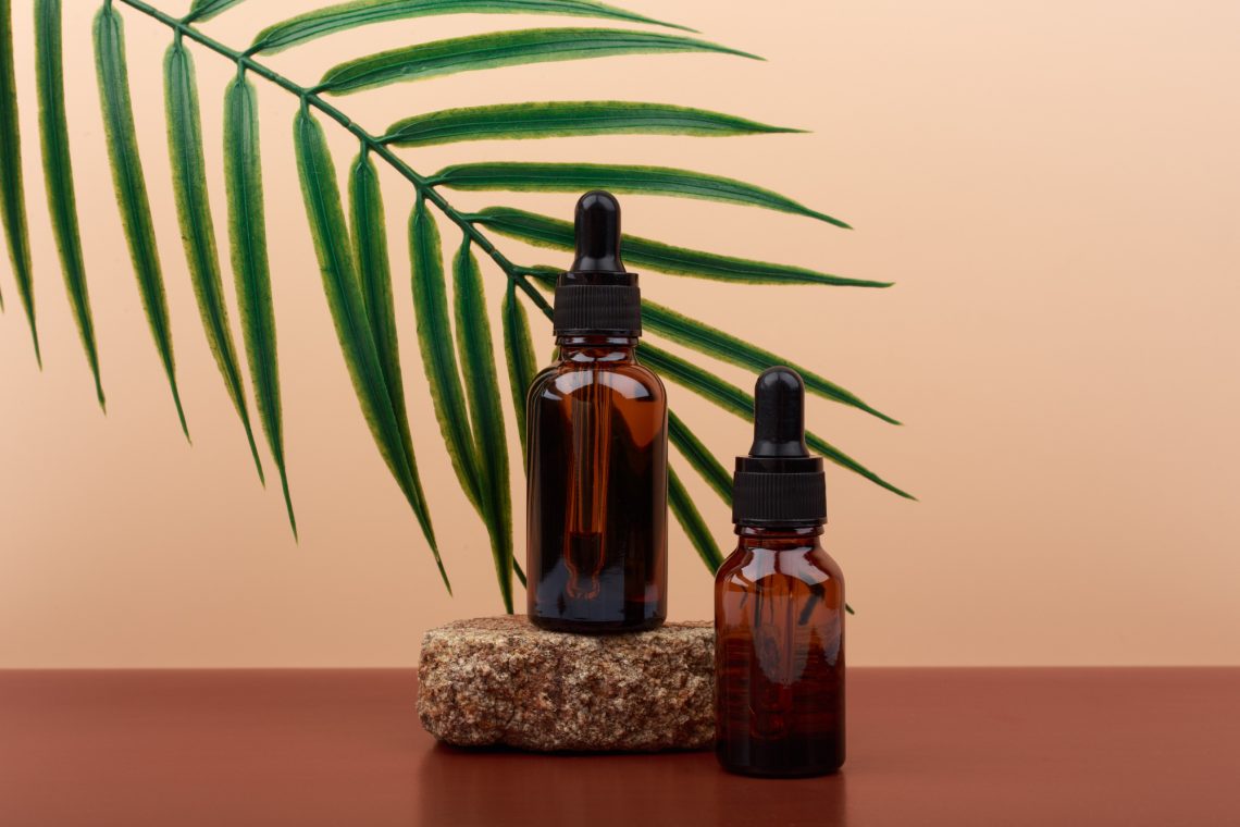 Two skin serums in dark glass bottles with black caps on brown stone against beige background with palm leaf
