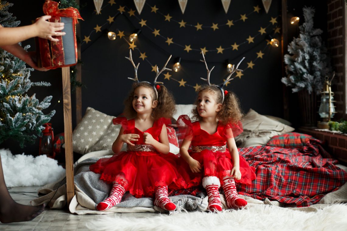 Young mother and her two little twin daughters opening a Christmas gift by a Christmas tree in cozy living room in winter. Girls in red dresses with deerhorns on head