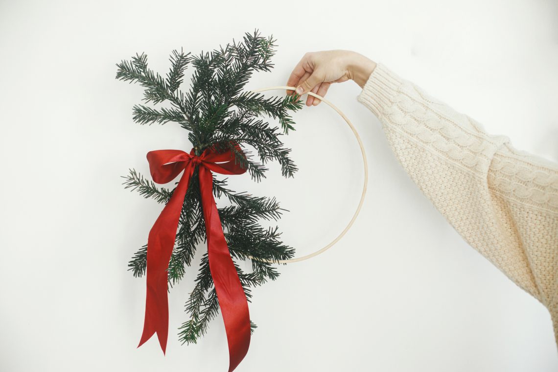 Hand in cozy sweater holding modern christmas wreath on white wa