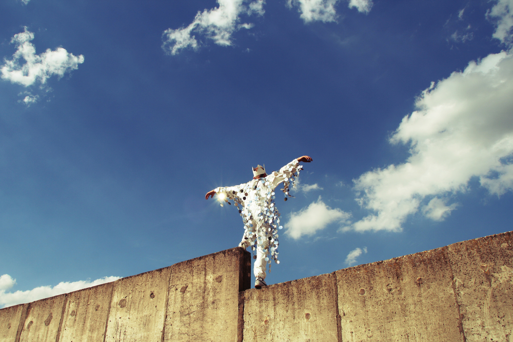 a man standing on a fence covered with bright pieces of decoration with sky as a backdrop