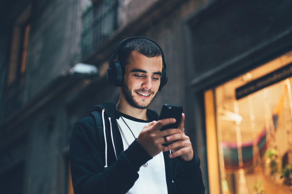 Фото молодий чоловік у чорному худі і великих бездротових навушниках посміхається, дивлячись на екран телефону