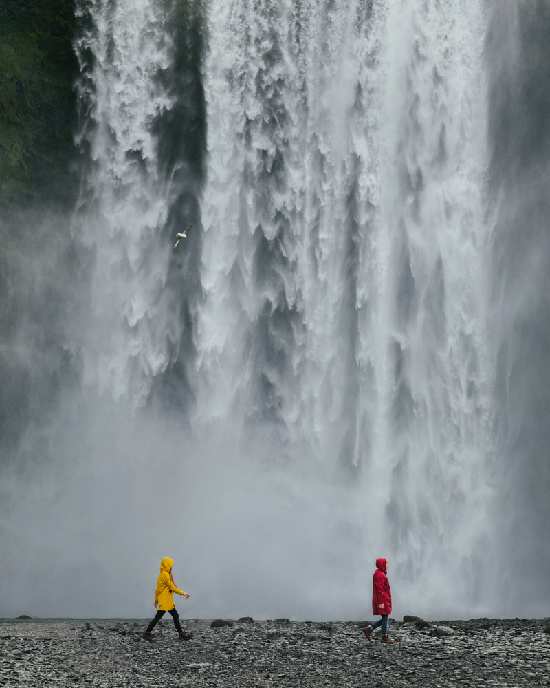 Couple of travelers in colored raincoats on the background of a large waterfall Skogafoss in Iceland. Epic landscape travel active tourism and adventure content.