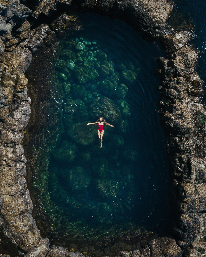 Beautiful girl in a red swimsuit swims in a round natural pool. Top view of a drone on a girl in a pool in Iceland on the Atlantic Ocean. Blue clear water.