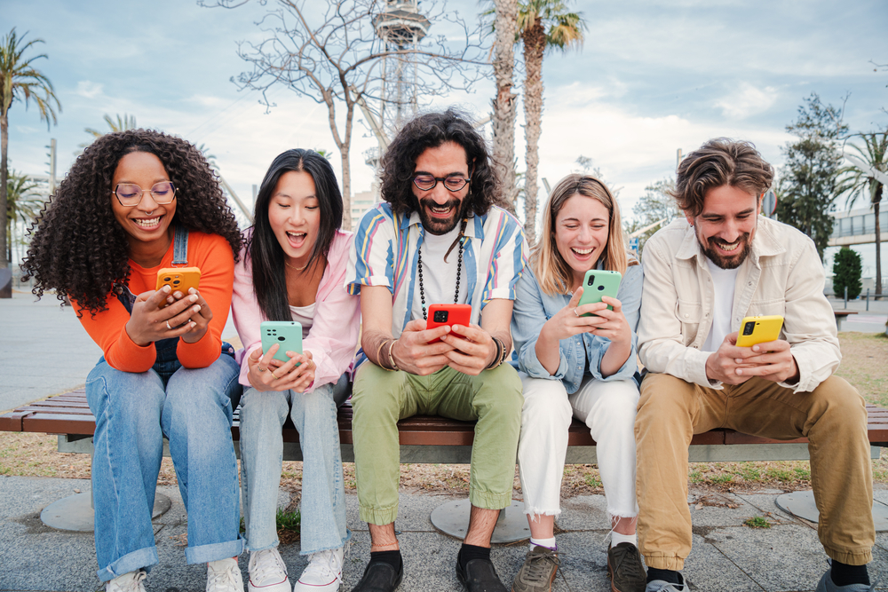 Photo Group of best friends enjoying and smiling using their mobile phone app sitting on a bench sharing messages. Happy young people chatting on social media online with the cellphone having fun together