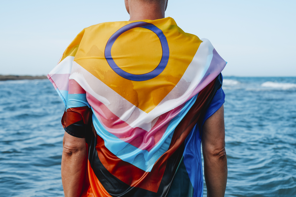 Photo closeup of a wet person in the sea, standing in the water, wrapped in an intersex-inclusive progress pride flag, seen from behind